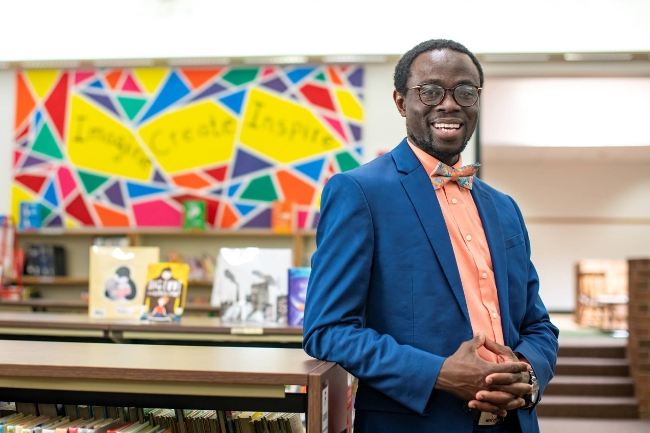 Black man swearing a bow-tie and blazer smiling at a counter in a library